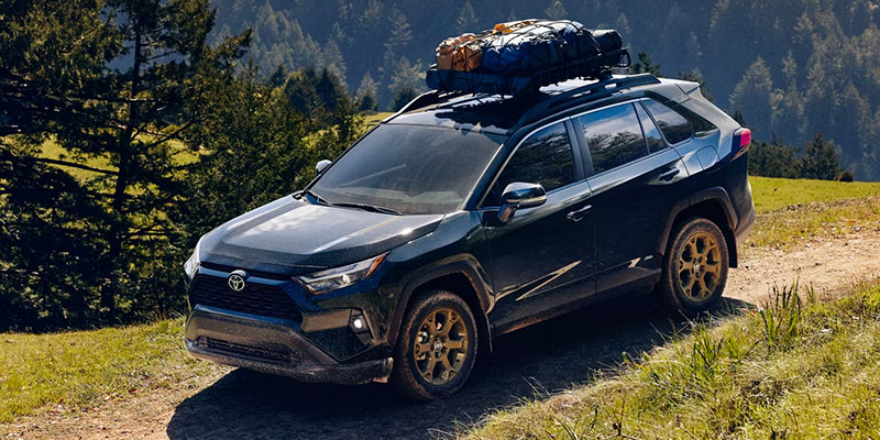 A dark green 2025 Toyota RAV4 driving on a scenic dirt trail with camping gear secured on the roof.