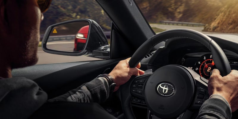 Driver gripping the steering wheel of a 2025 Toyota GR Supra , showing part of the dashboard and side mirror