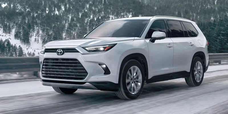 Front view of a white 2025 Toyota Grand Highlander driving through snowy forest roads