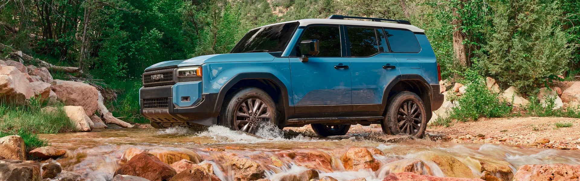 2025 Toyota Land Cruiser crossing a shallow creek in a rocky forest setting.